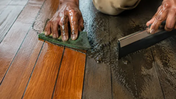 A green pad and rubber squeegee lifting dark oily slurry off a soiled timber floor, revealing clean warm timber behind