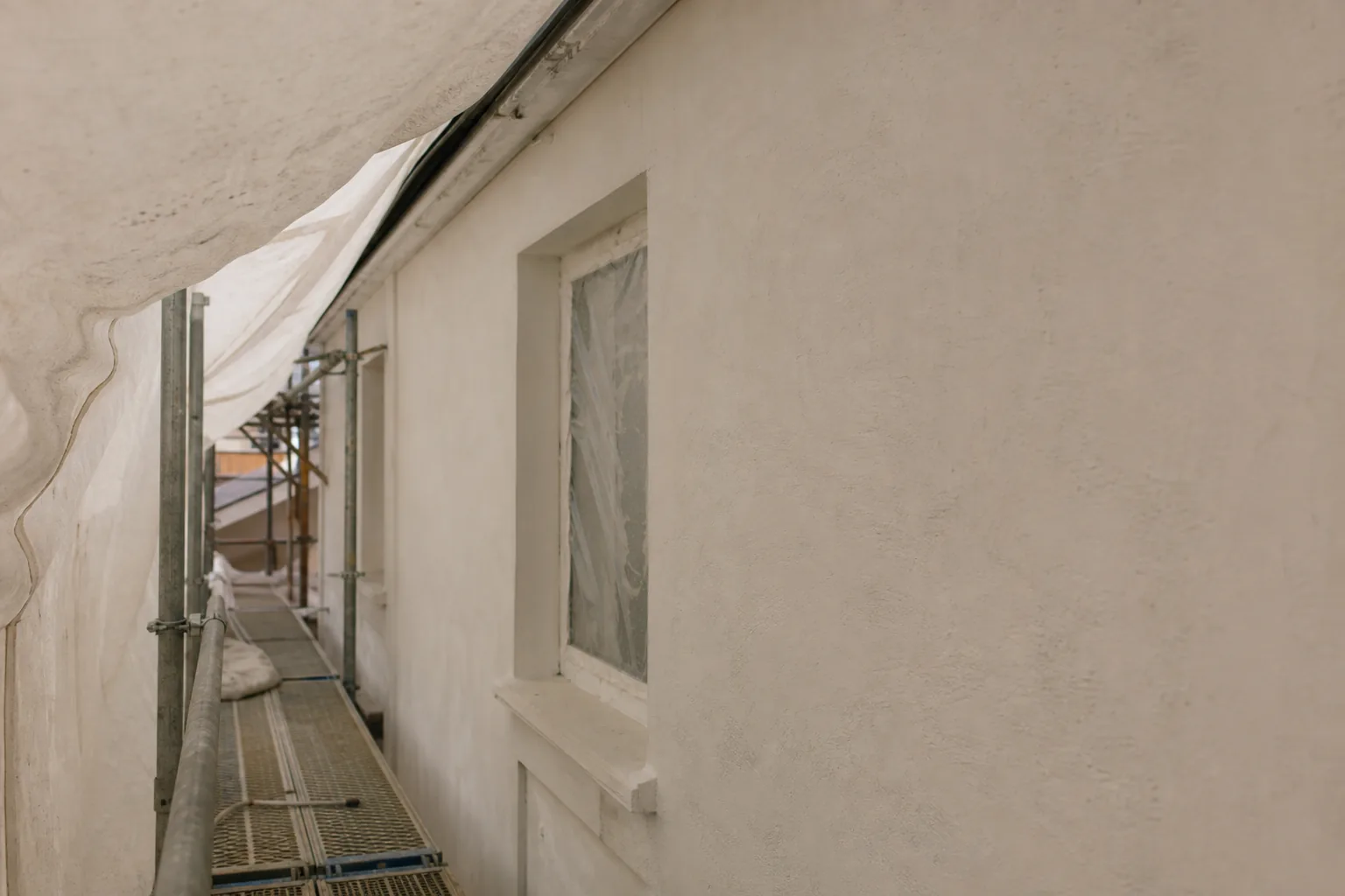 A freshly primed matte off-white plaster facade wall drying under scaffolding and protective sheeting, shielded from sun and rain