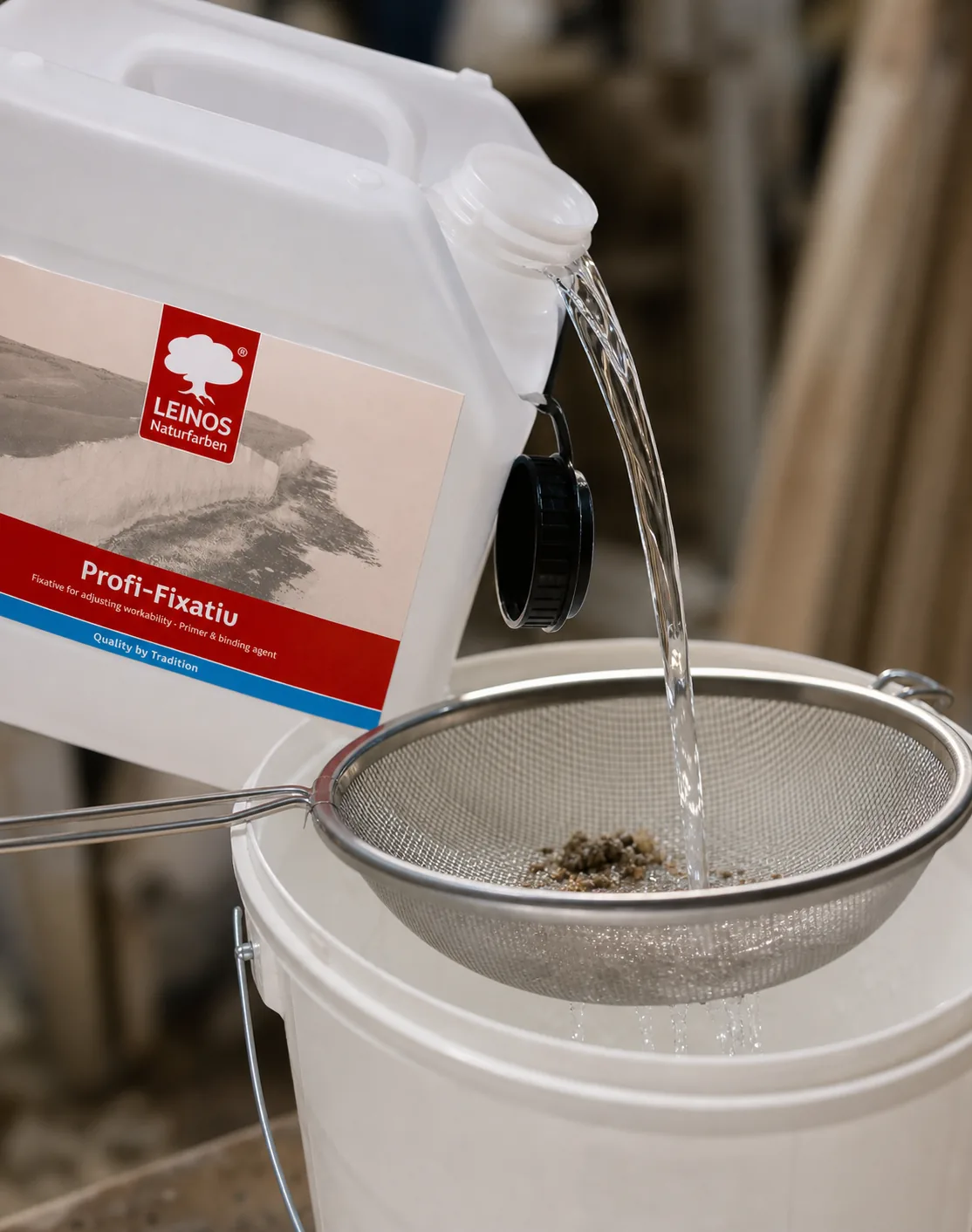 Clear water-glass primer being poured through a fine sieve from the branded LEINOS Profi-Fixativ white canister into a clean bucket