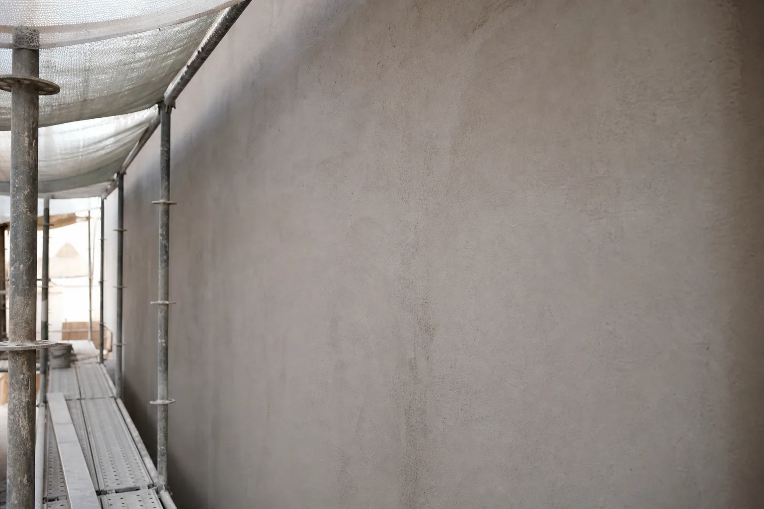 A freshly primed matte raw plaster facade wall firming under scaffolding and protective sheeting, shielded from sun and rain