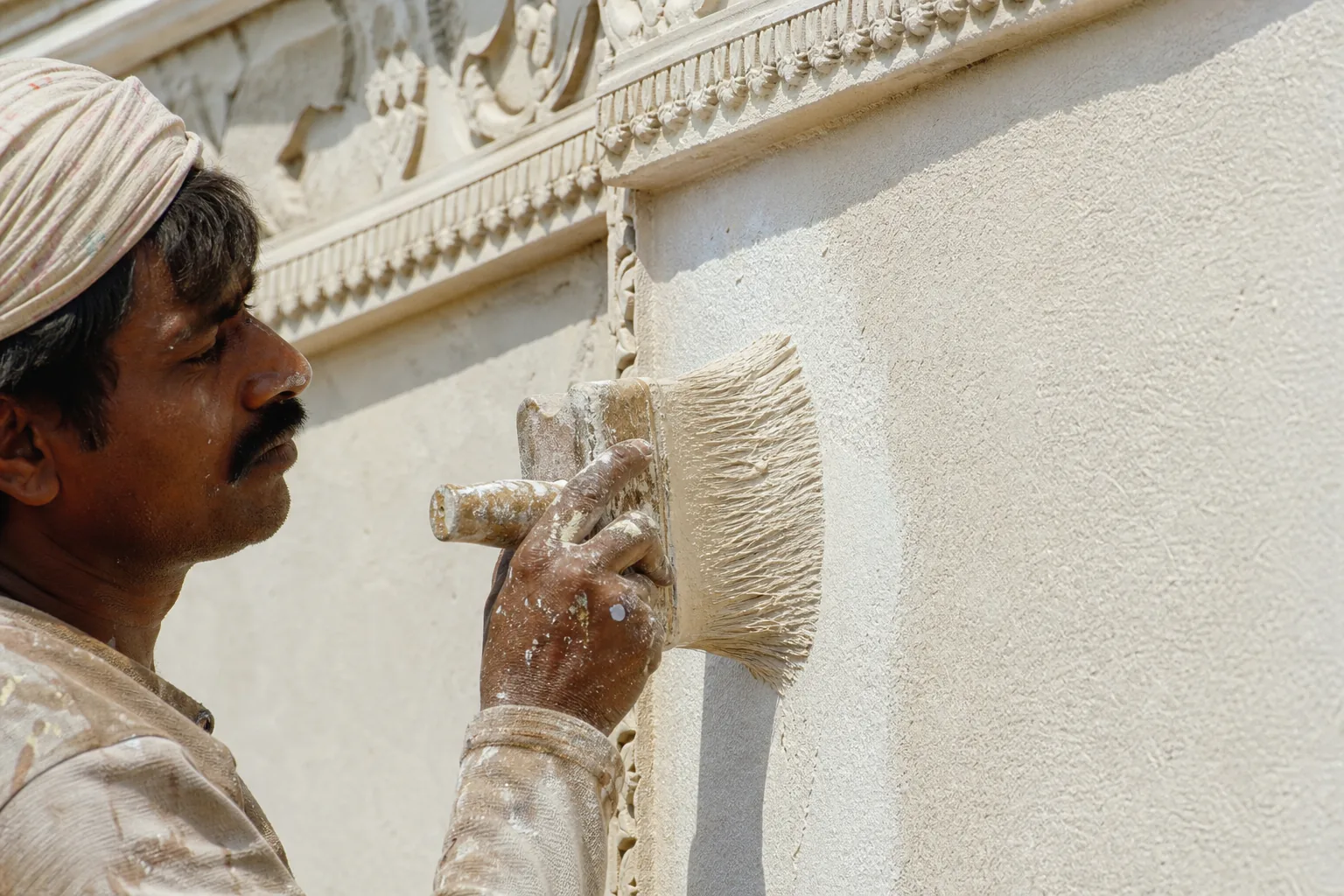 Indian craftsman brushing a thick coarse off-white mineral ground coat full-surface onto an exterior plaster facade with a wide block brush
