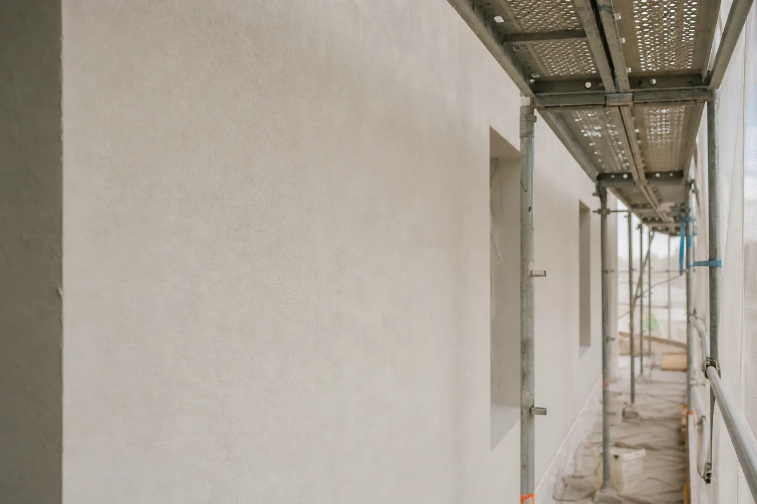 A freshly primed matte off-white plaster facade wall with a subtle grainy texture drying under scaffolding and protective sheeting