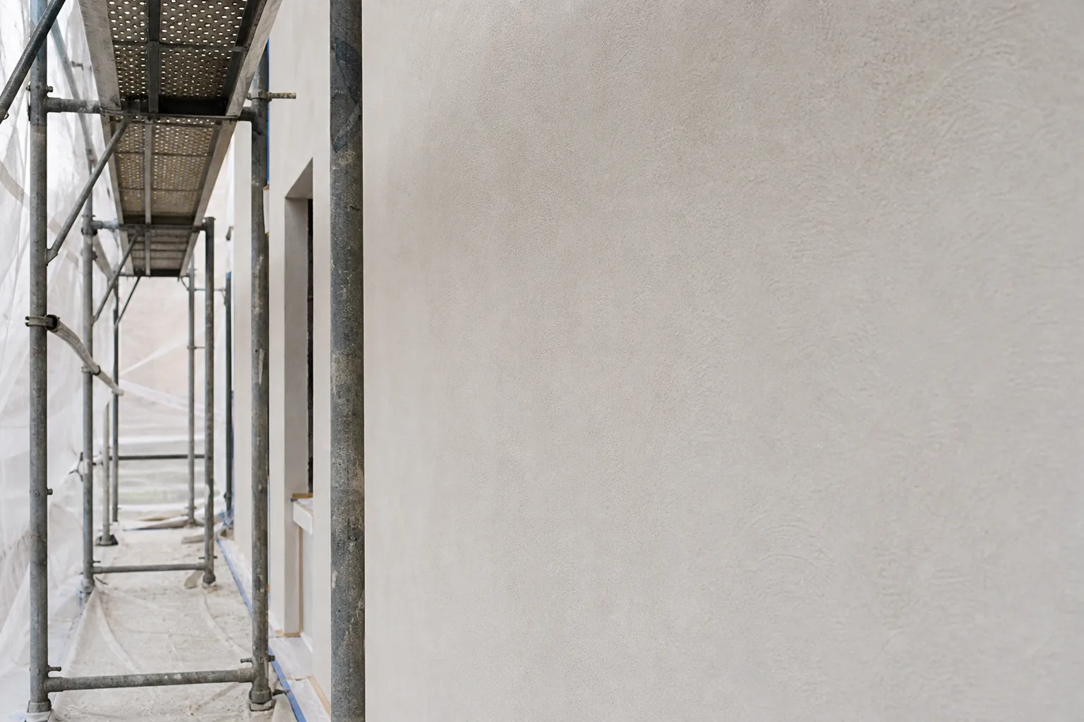 A freshly primed matte off-white plaster facade wall with a subtle grainy texture drying under scaffolding and protective sheeting