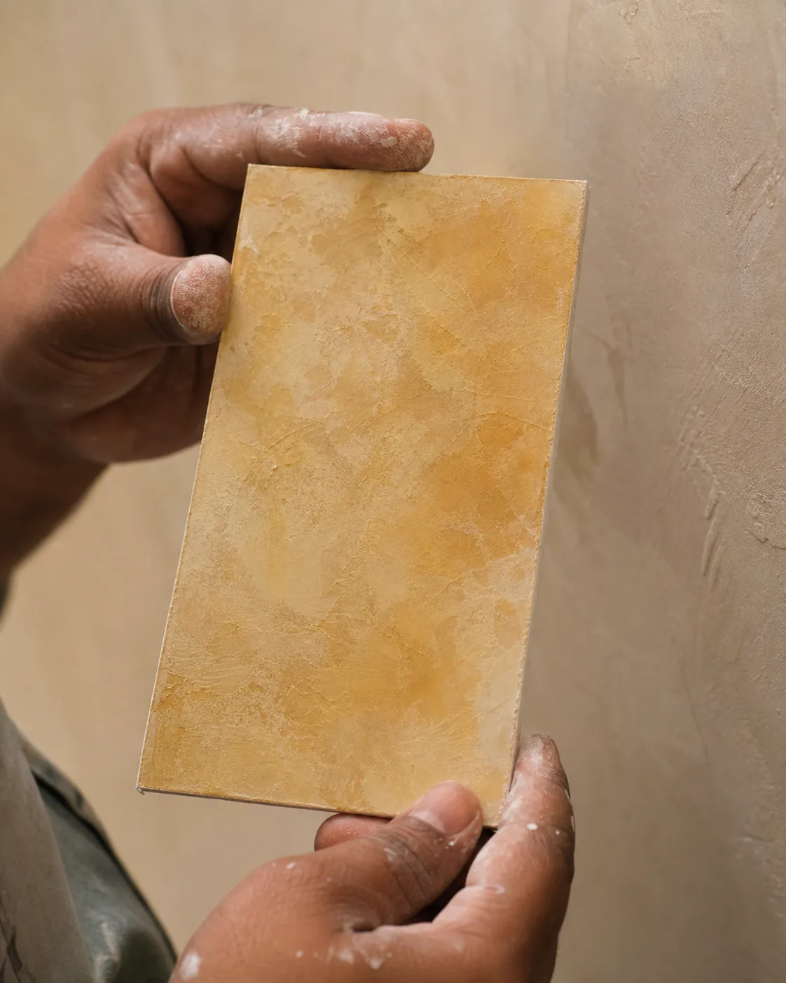 Indian decorator’s hands holding a coated sample board with a warm translucent scumble-plaster test patch against an interior wall