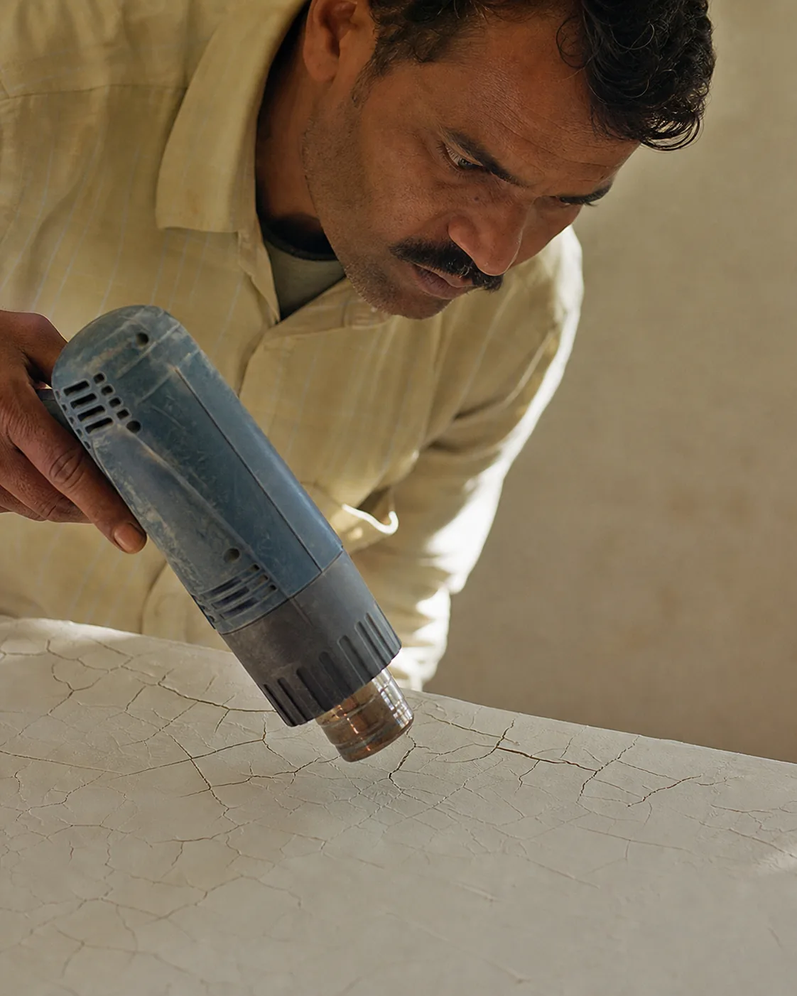 Indian craftsman working a heat gun in circular motions over a freshly spatulaed wall, fine tension cracks forming