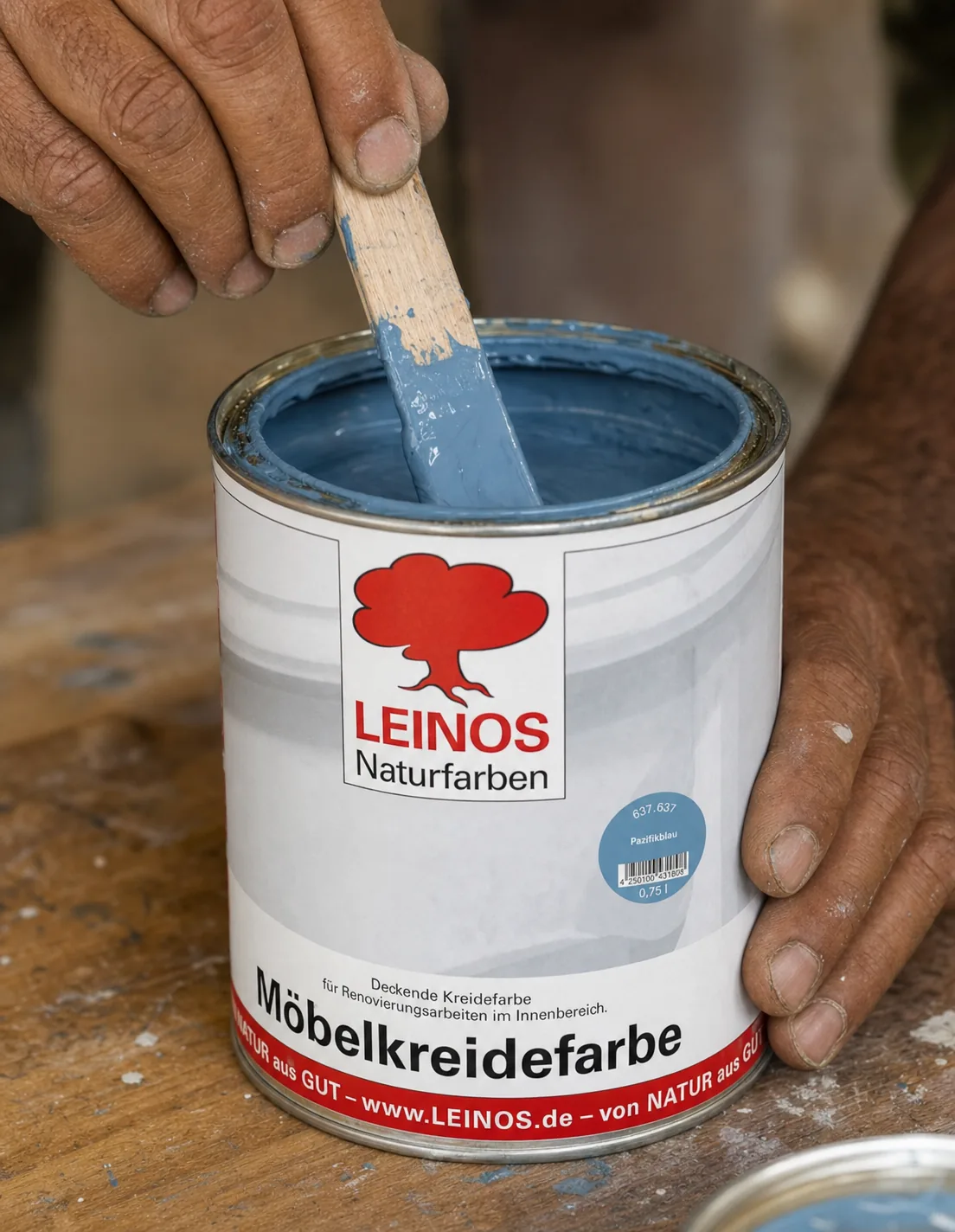 Indian craftsman stirring matt Pacific-blue chalk paint inside an open branded LEINOS Furniture Chalk Paint tin with a flat wooden stir stick on a workbench