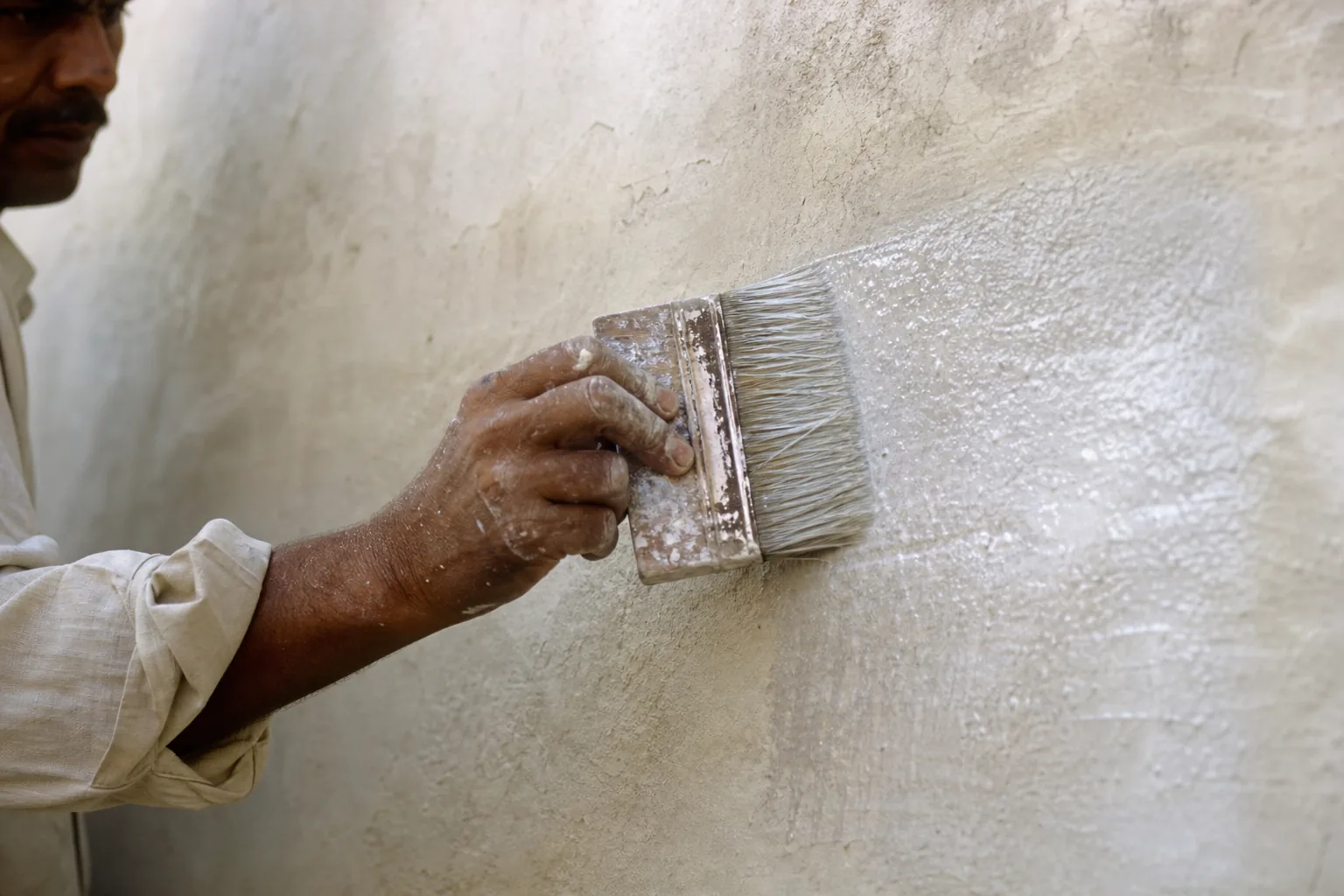 Indian craftsman brushing whitish mineral primer onto a patch of chalking lime plaster wall with a flat masonry brush