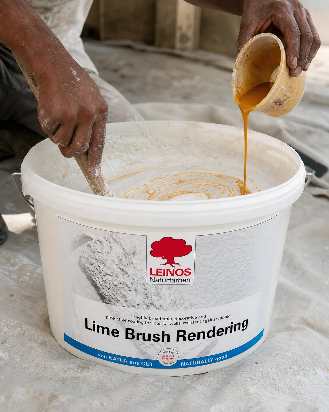 Indian craftsman stirring thick creamy white lime render in an open LEINOS bucket with a wooden stick, a measured pour of ochre pigment concentrate being added