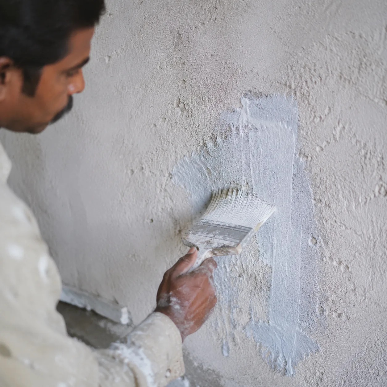 Indian craftsman brushing whitish mineral primer onto a patch of chalking absorbent plaster wall with a flat masonry brush
