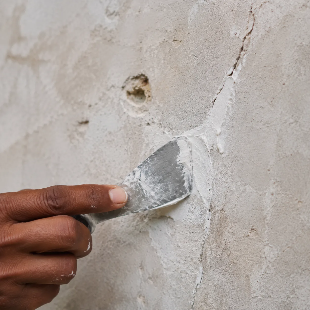 Indian craftsman pressing bright-white lime filler into a wall crack with a stainless filling knife, pulling it flat into a thin layer