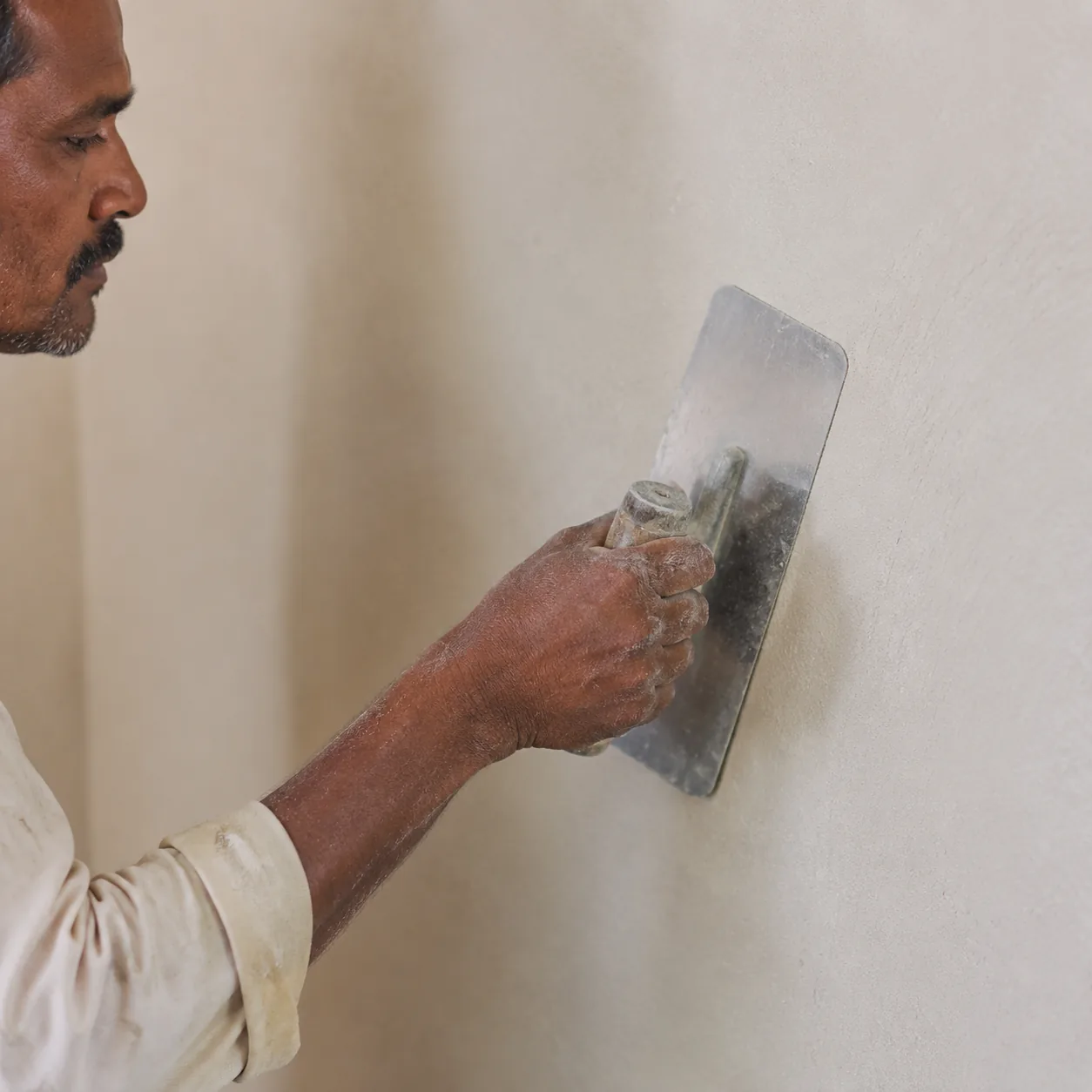 Indian craftsman smoothing and felting a filled interior wall flat with a wide stainless trowel, an even matte off-white lime skim building up