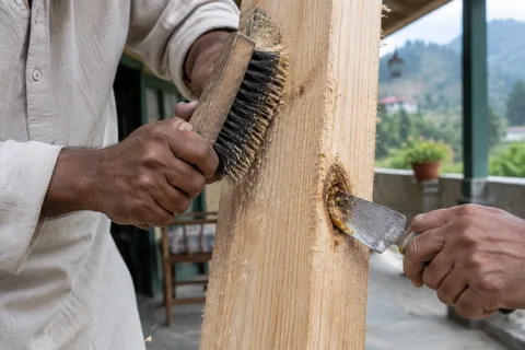 A craftsman scrubbing rough-sawn pine cladding with a stiff brush and scraping out a resin pocket before painting