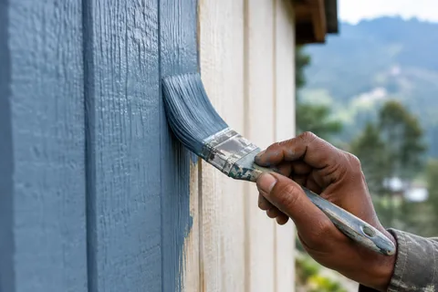A flat brush laying a thin first coat of Frisian blue weatherproof paint along rough-sawn cladding boards
