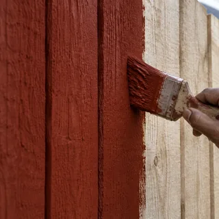 A brush applying matt Swedish red water-based paint to untreated rough-sawn fence boards