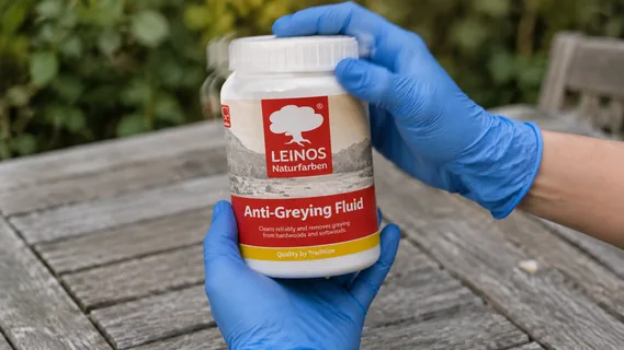 Gloved hands shaking an upright jar of LEINOS Anti-Greying Fluid above a greyed teak garden table
