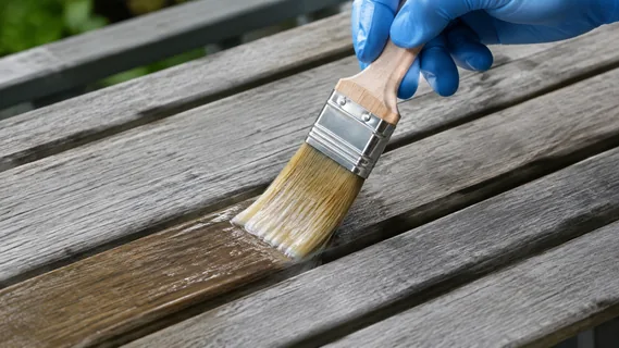 A gloved hand brushing anti-greying fluid undiluted onto silver-grey teak garden furniture slats