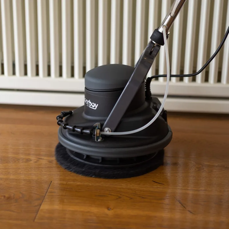 The Floorboy working a black pad into a timber floor beside a radiator