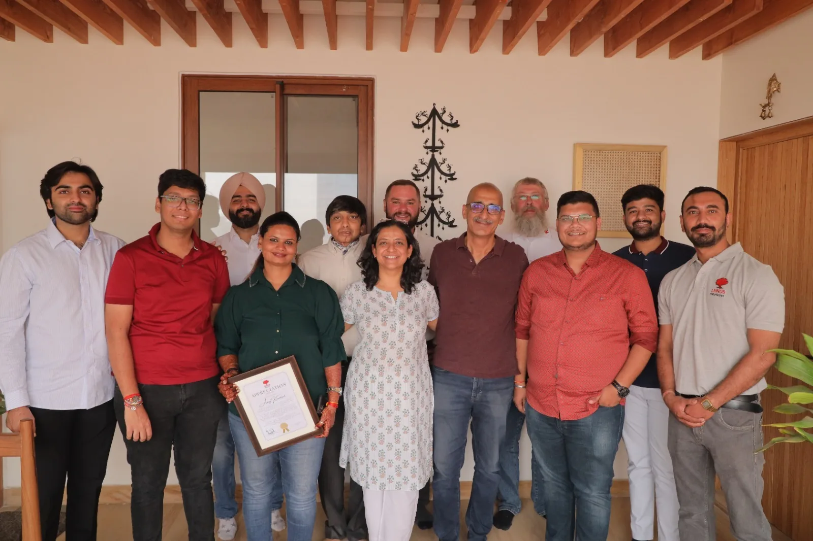 The full LEINOS India team and the client family gathered beneath the project's exposed wooden ceiling beams, with the framed LEINOS Appreciation certificate at the finished Noida pine bedroom.