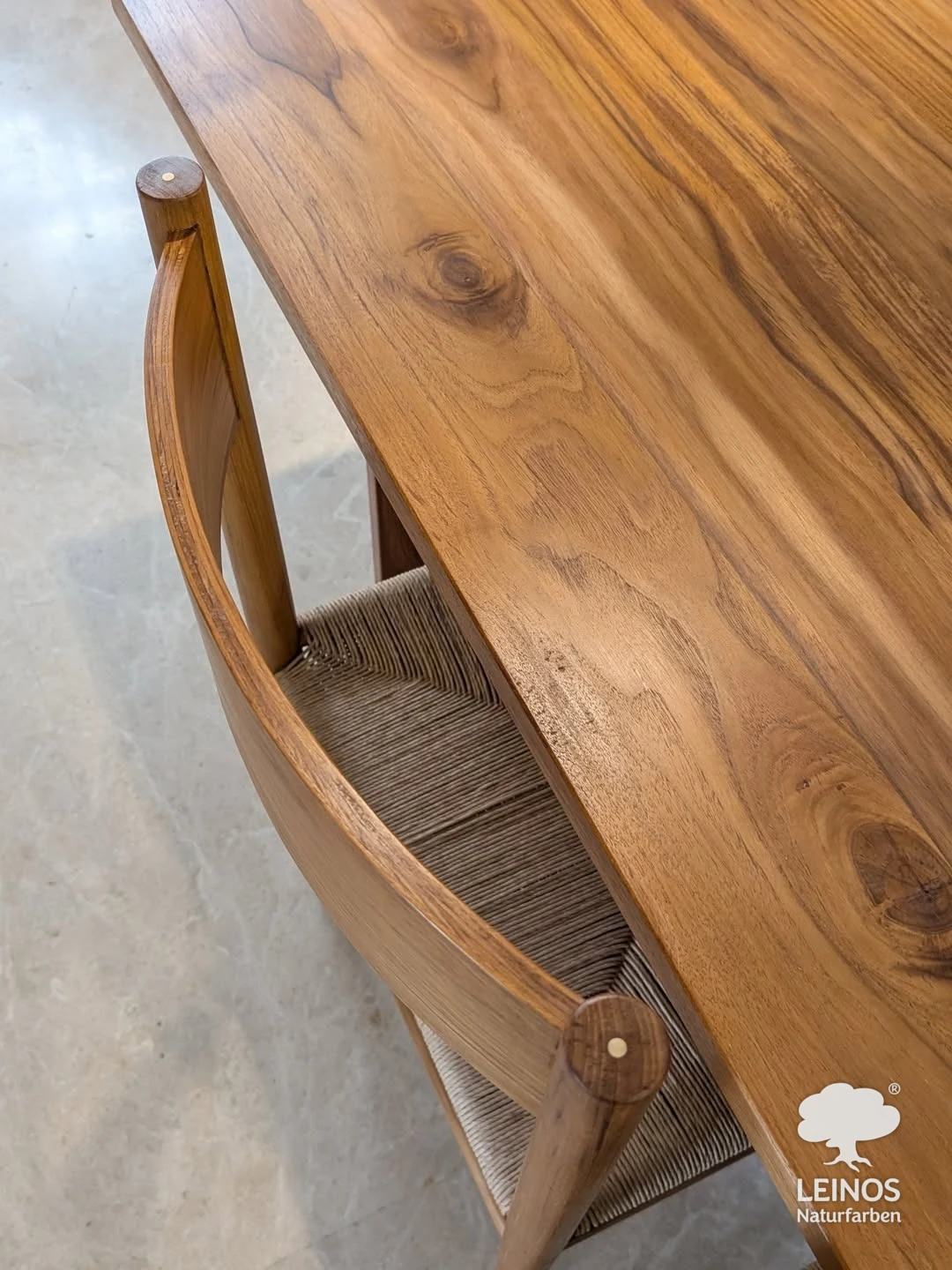 Close-up of the teak tabletop edge and a rush-seat dining chair — hardwax-oiled teak grain detail with brass-pegged joinery.