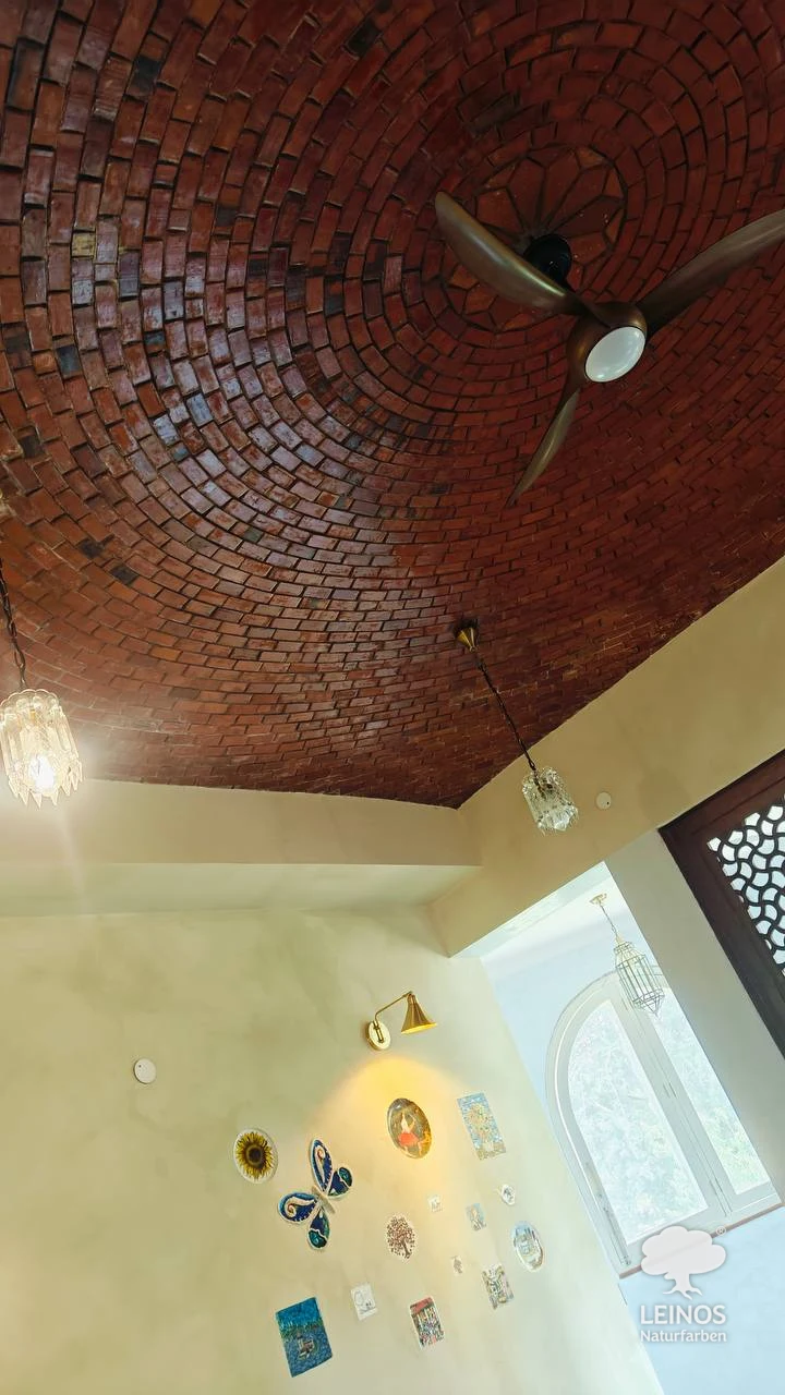 Looking up in the New Delhi interior — the exposed-brick spiral vault sealed in Stone & Concrete Oil 254 meets limewashed walls in Lime Paint 665, with a curated gallery of ceramics, an arched window and crystal pendants below.