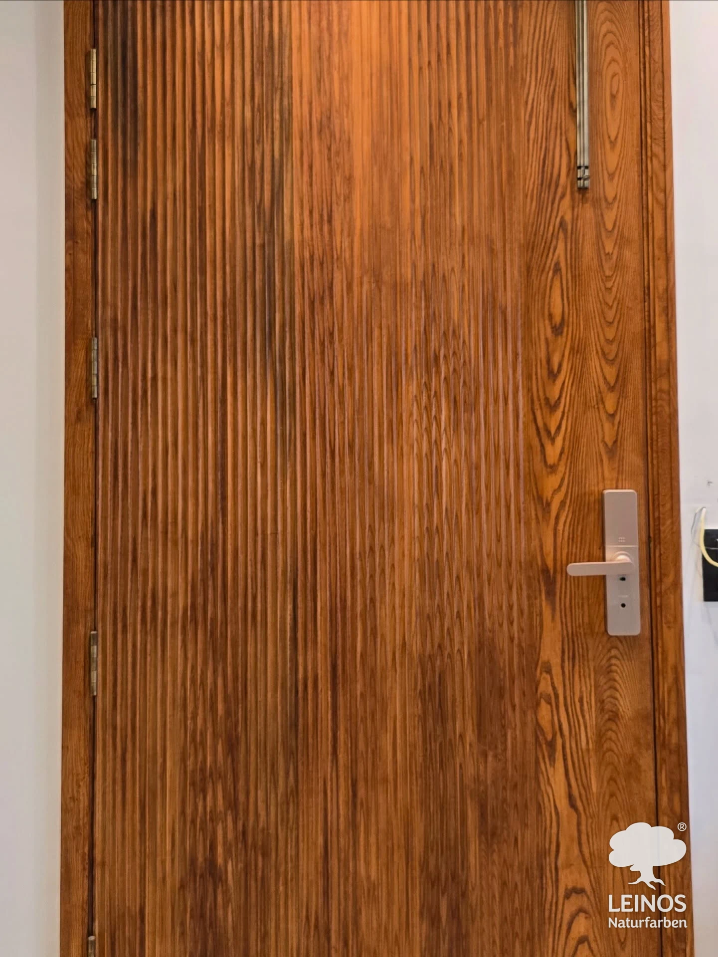 A reeded wood interior door in the same home, finished in Premium Hardwax Oil 290 — the open-pore oil keeps the vertical fluting and grain visible, paired with a brushed-metal smart lock and pull handle.