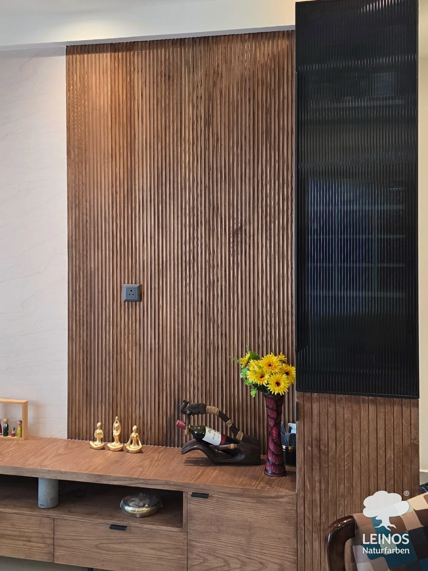 A closer view of the living-room fluted wood panelling and console in Premium Hardwax Oil 290, beside a dark reeded-glass cabinet and a vase of sunflowers.