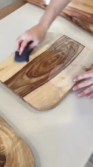 An @leinos.india Reel: a maker in Bengaluru hand-rubs LEINOS Countertop Oil 280 into a figured-wood serving and chopping board with a cloth, the pale-and-dark grain deepening under the oil. A close-up finishing demo of a food-contact-safe penetrating oil (WESSLING-tested to EC 1935/2004 + LFGB), not a full project walkthrough.