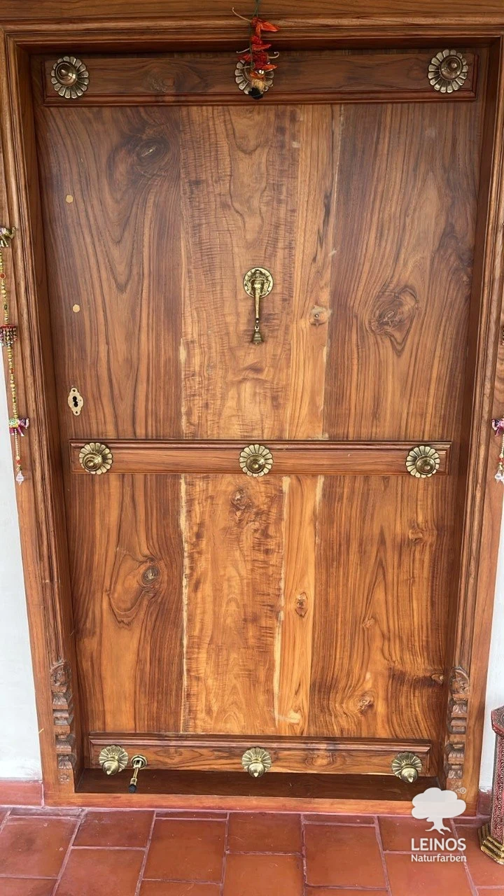 A carved teak entrance door with brass rosettes and a bell pull in a private Bengaluru home, finished in LEINOS Interior Hardwax Oil 290 — open-pore natural teak grain, no film.