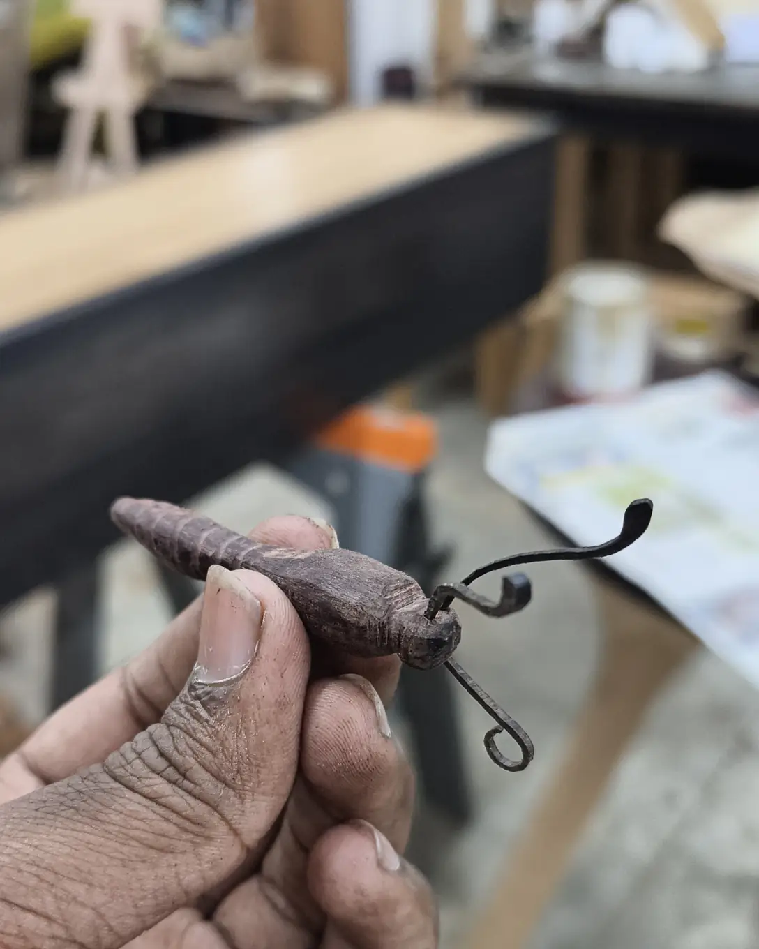 Fingers hold the carved rosewood body of the butterfly, with both antennae curled at the tip, in front of a workbench.