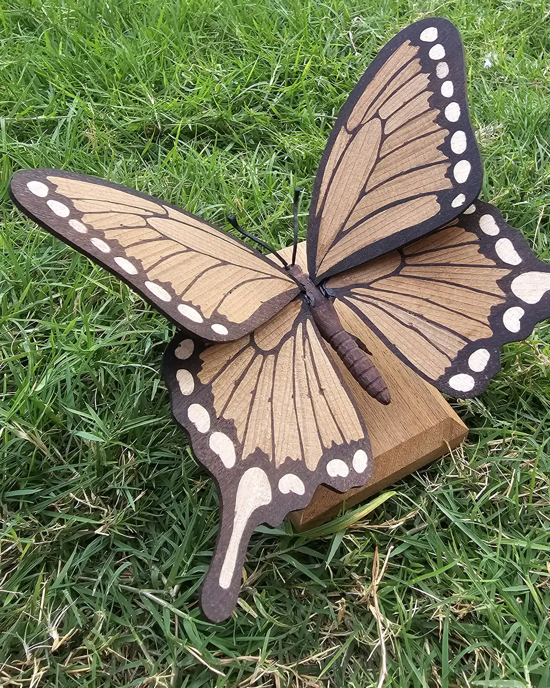 A wooden marquetry butterfly with raised ochre wings and a dark ebony border stands on a small teak block on cut grass.