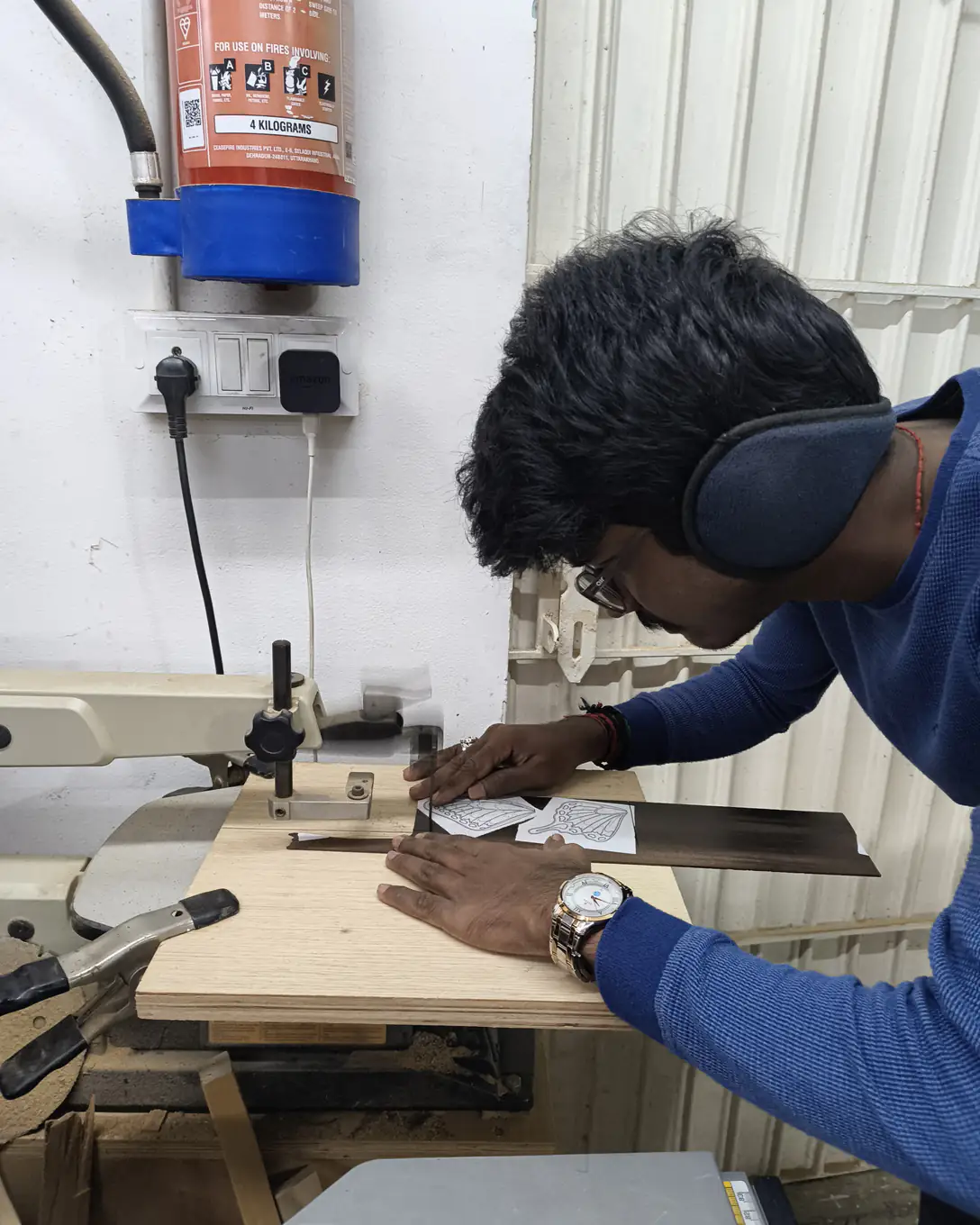 Haris in ear defenders bends over a scroll saw in the studio workshop, cutting a small veneer piece with a paper pattern taped to it.